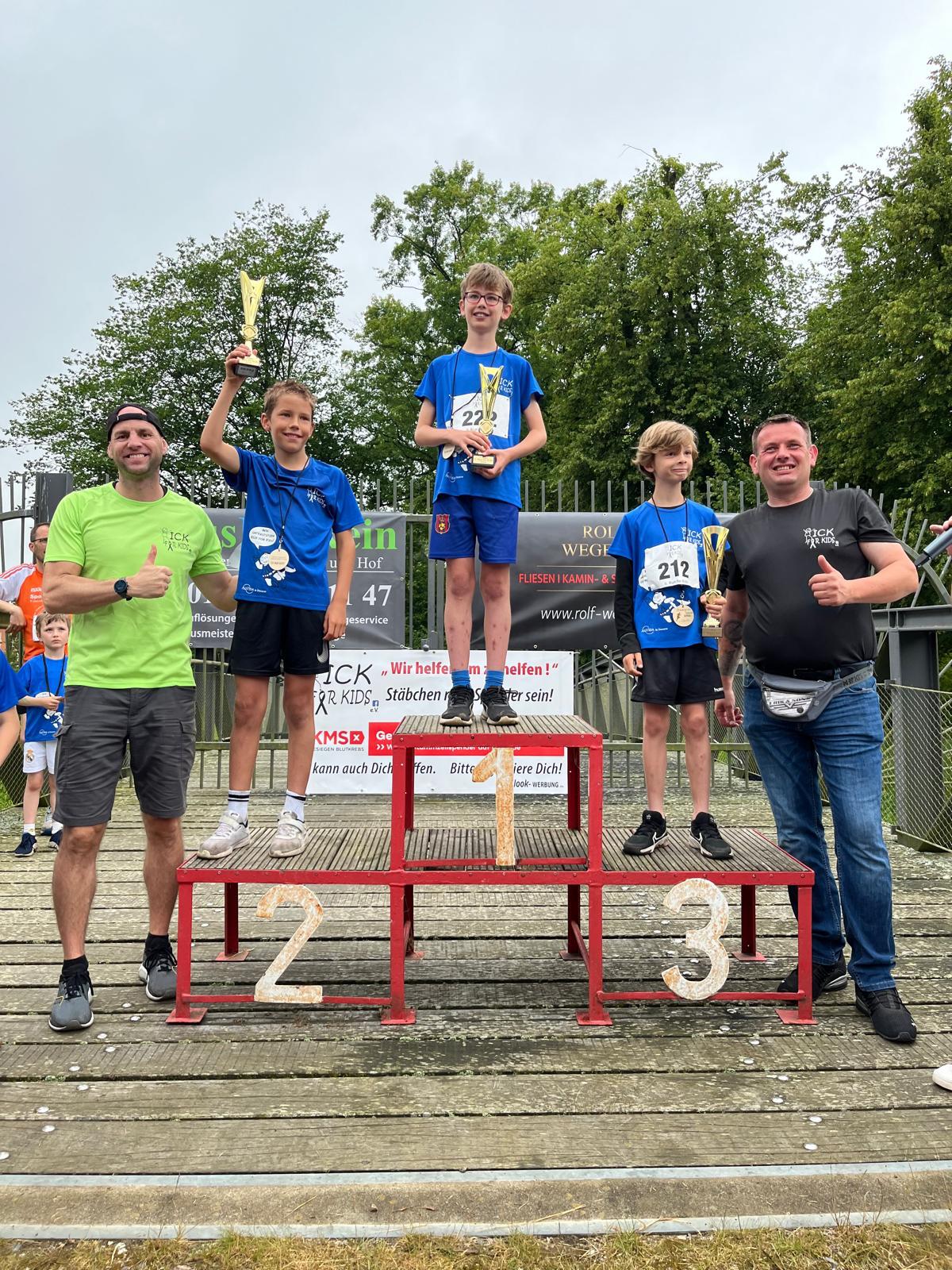 Siegerehrung beim Kinderlauf: drei Kinder auf dem Treppchen mit Pokalen, flankiert von zwei Betreuern